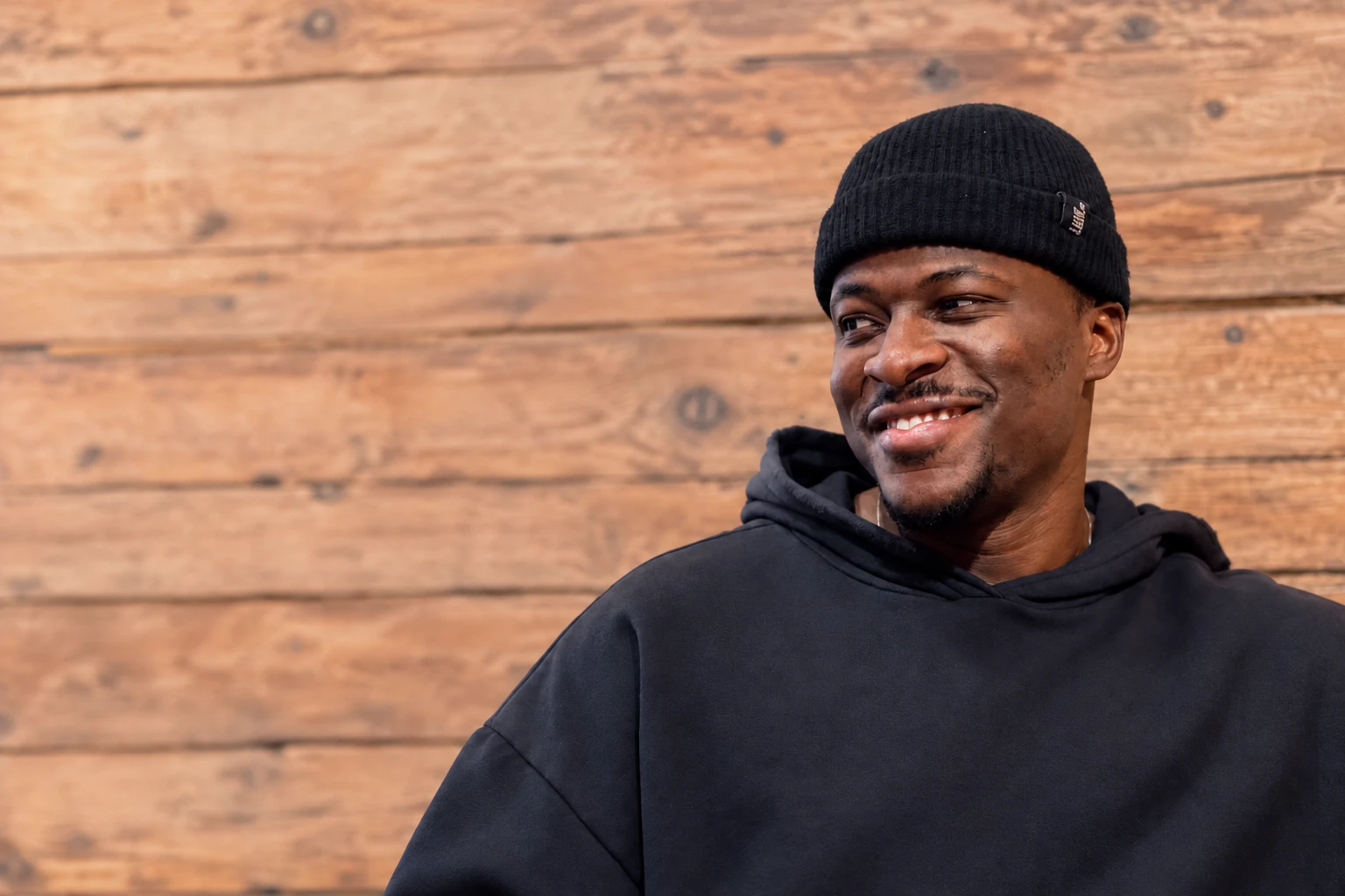 Olu Roberts smiling in a black beanie and dark hoodie against a log wall